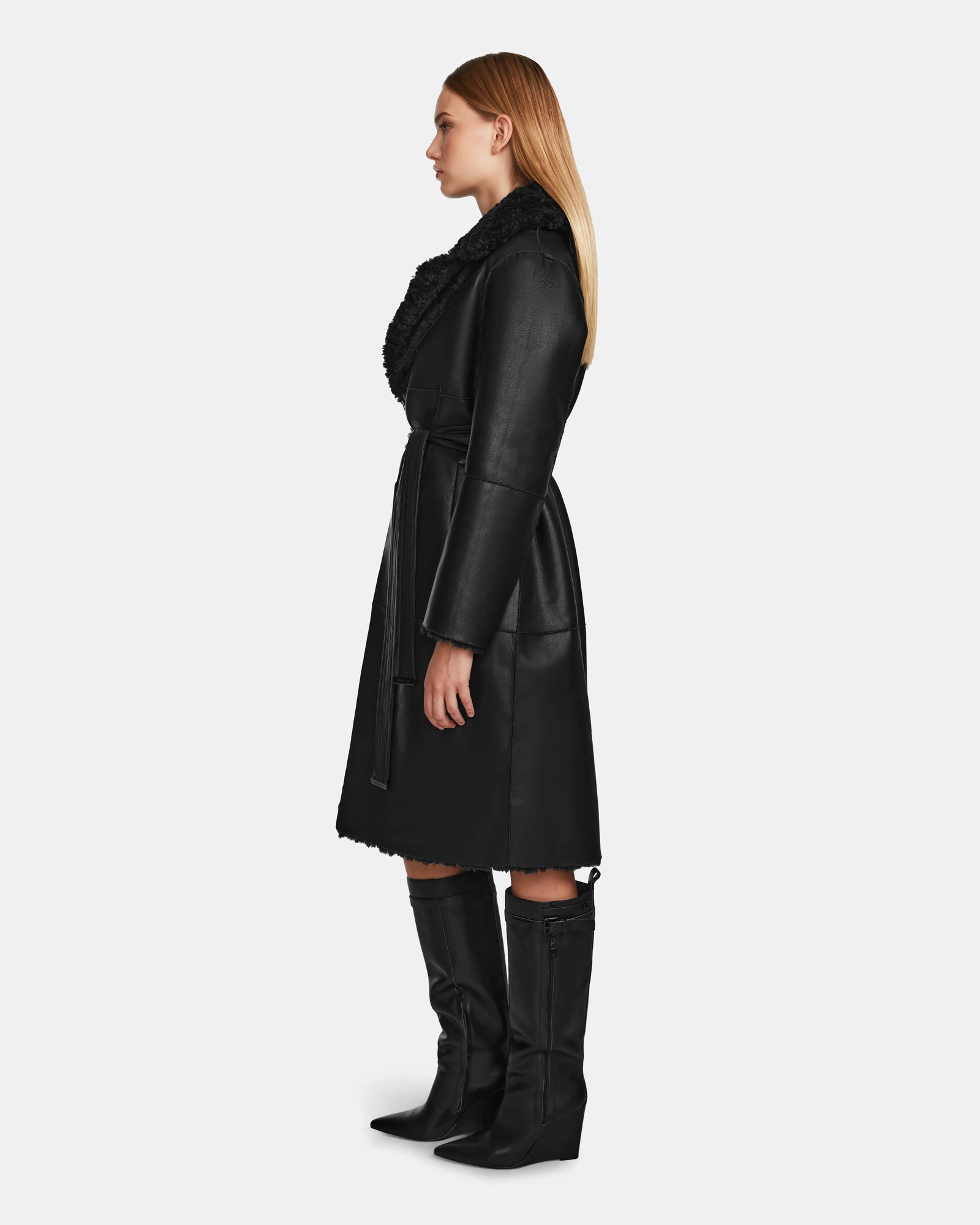 A woman with straight blonde hair stands in profile, wearing the Rudsak ANNABELLE - L124544 Black vegan leather coat with a textured collar and knee-high black boots, posed against a plain white background.
