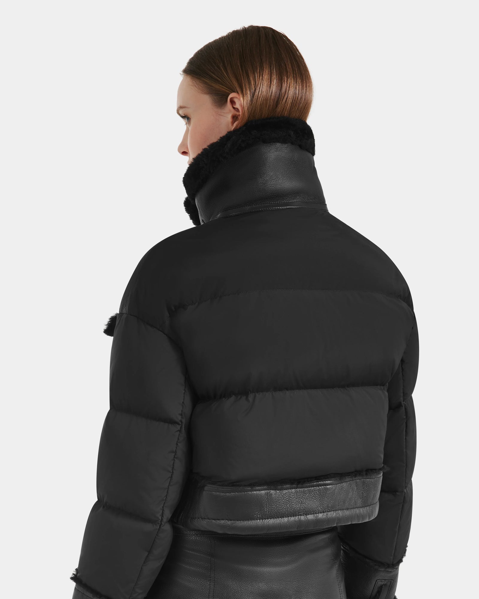 A person with straight brown hair models the Rudsak ARIELLA - 8125505 Black women’s cropped shearling puffer, featuring a high collar and leather accents, against a plain light background—perfect for cold-weather styling.