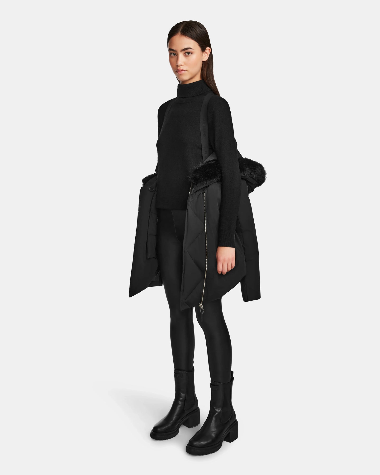 A woman stands facing forward in a black turtleneck, black leggings, and boots, holding the Rudsak AURORE - L123561 Black/Black women’s short fitted down parka with fur-trimmed hood over her arm against a plain white background.