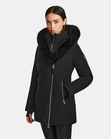 A woman models the Rudsak AURORE - L123561 Black/Black jacket, featuring a fitted design, large fur-trimmed hood, silver zippers, and a sleek modern look, standing with one hand in her pocket against a white background.