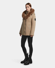 A woman wears the Rudsak AURORE - L123561 Sand fitted winter coat, featuring faux fur trim, paired with black leggings and ankle boots, standing against a plain white background.