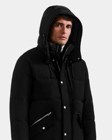 A man wearing the Rudsak BRADY - M125510 Black men's parka with a removable hood, zipped over a dark layer, stands against a plain light background, gazing slightly to the side.