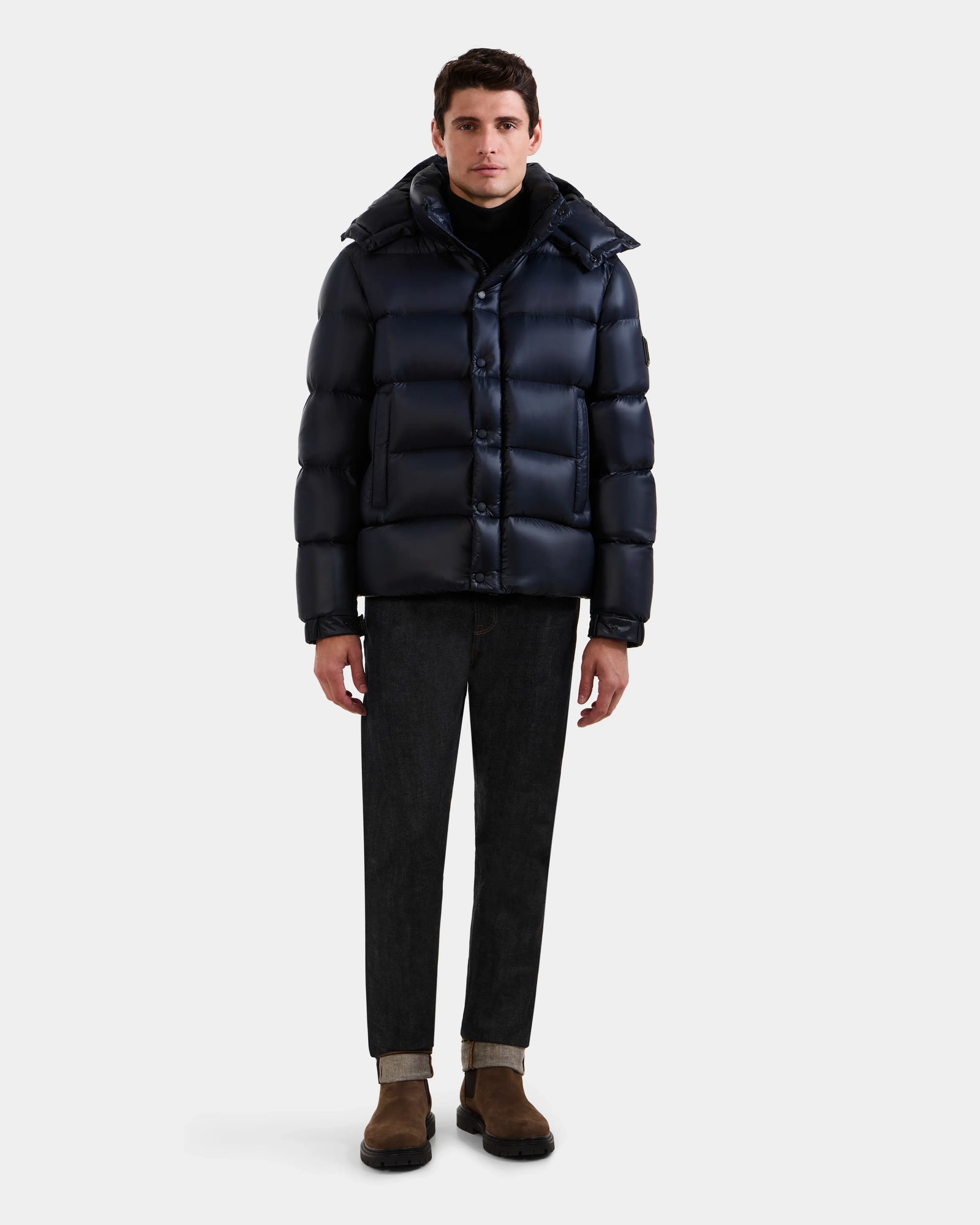 A man faces forward in a Rudsak BRAYDEN S - M125525 Navy jacket, black turtleneck, dark cuffed jeans, and brown suede boots against a plain white background.