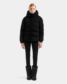 A person faces forward wearing the Rudsak BREE M - L125582 Black women’s down puffer with removable hood, black leggings, and black winter boots against a plain light gray background.
