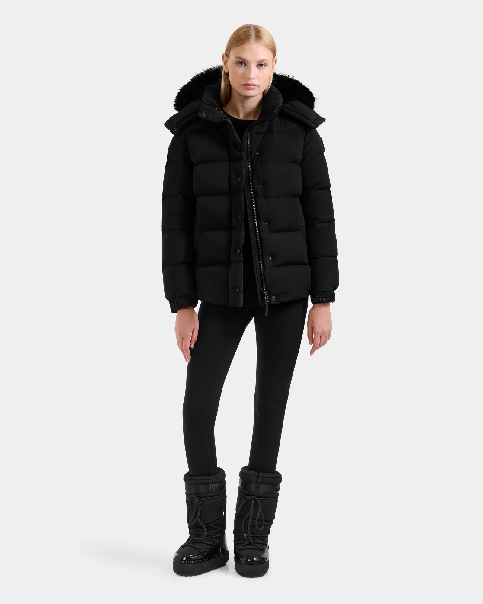 A person stands facing forward in the Rudsak BREE M - L125582 Black women’s down puffer jacket with a fur-trimmed removable hood, black leggings, and winter boots against a plain light background—a cold-weather essential.
