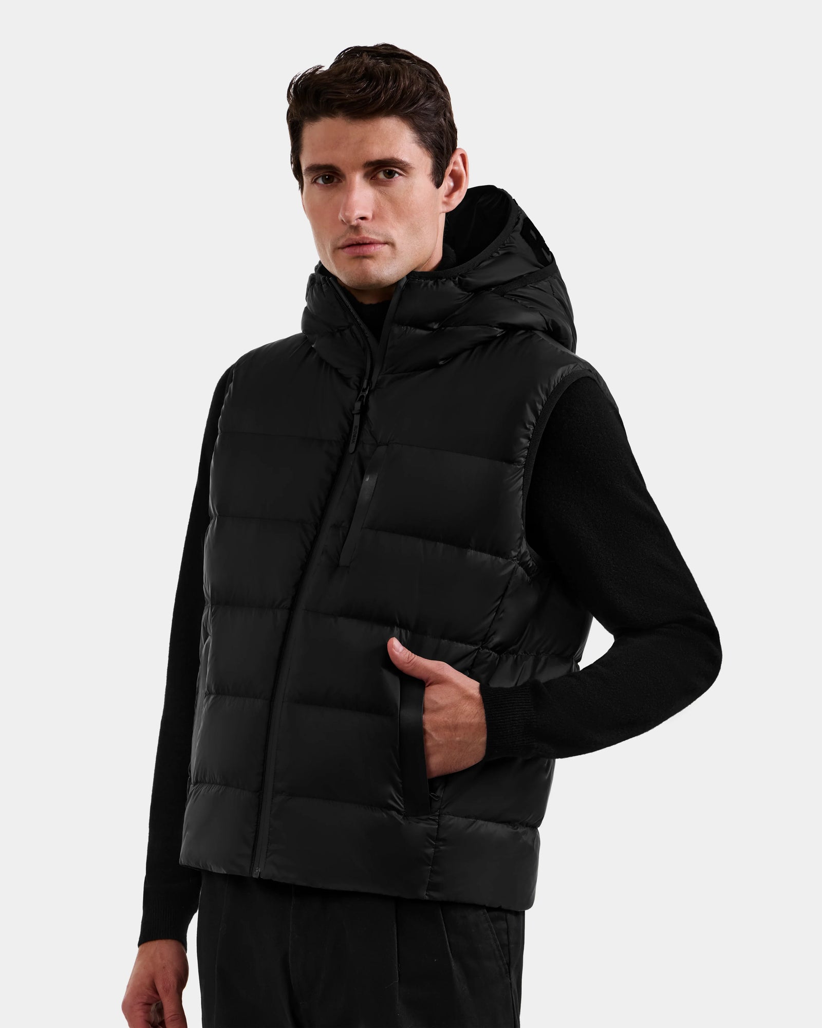 A man wearing a Rudsak BROOKS - M125520 Black men’s down puffer vest with a recycled nylon shell over a black long-sleeve shirt stands with one hand in his vest pocket, looking slightly to the side against a white background.