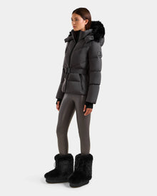A woman wears the Rudsak CHRISTELLE M - L125542 Grey quilted down puffer jacket with a belted waist and fur-trimmed hood, shiny gray leggings, and black fur boots against a plain white background.