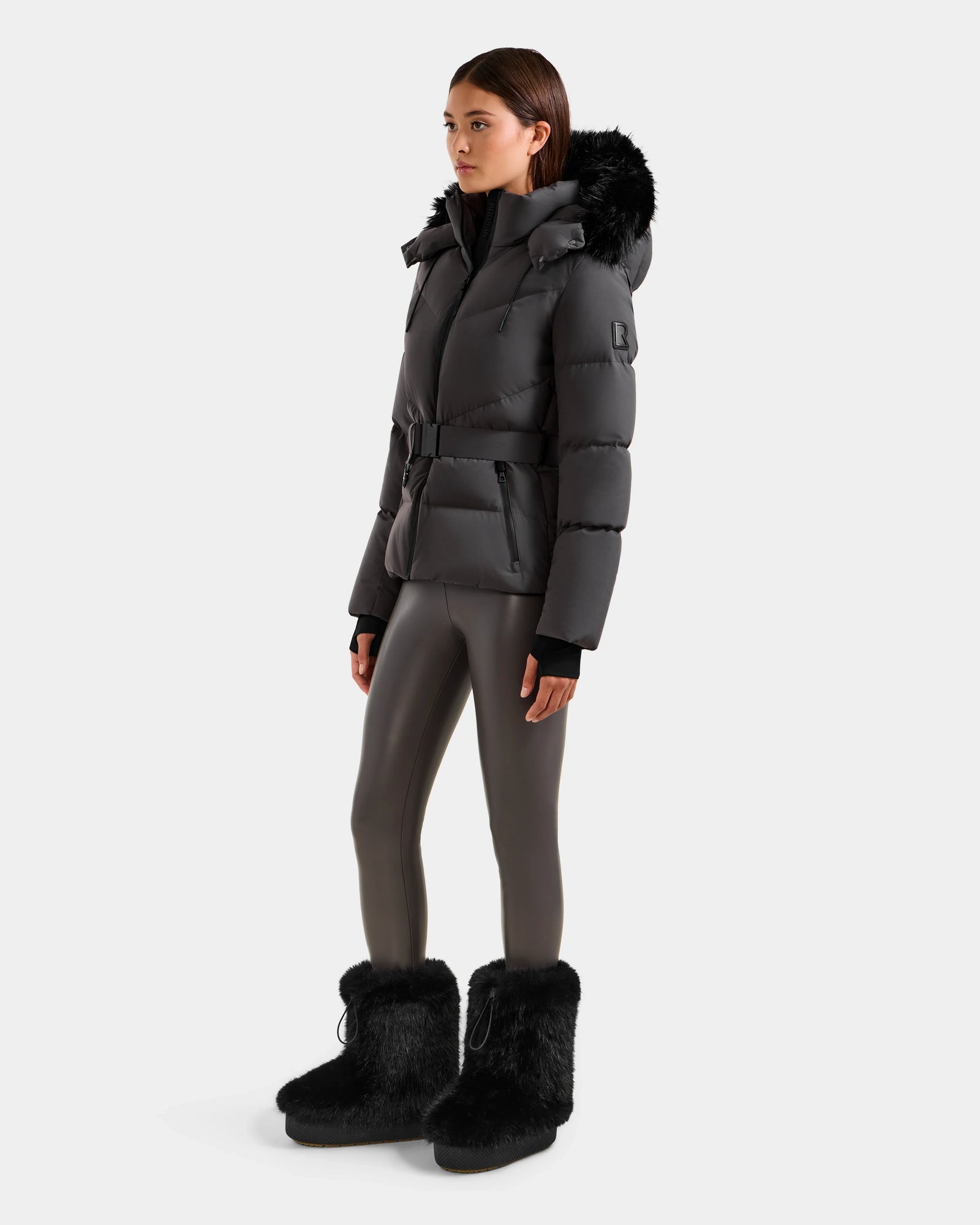 A woman wears the Rudsak CHRISTELLE M - L125542 Grey quilted down puffer jacket with a belted waist and fur-trimmed hood, shiny gray leggings, and black fur boots against a plain white background.