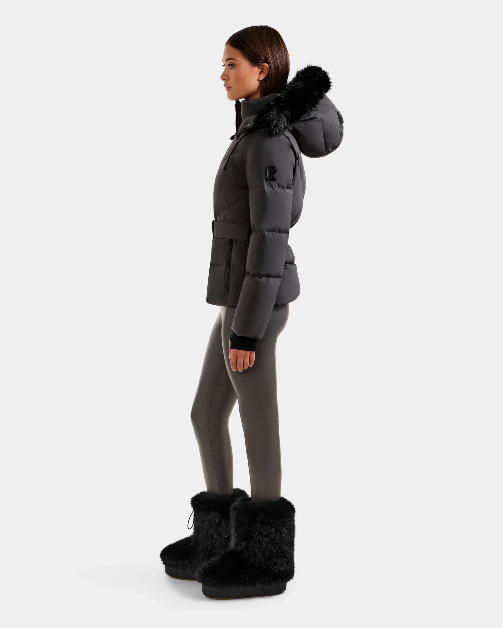 A woman stands in profile wearing the Rudsak CHRISTELLE M - L125542 Grey weather-resistant jacket, featuring a black puffer style with a fur-trimmed hood, paired with gray leggings and black furry boots against a plain white background.