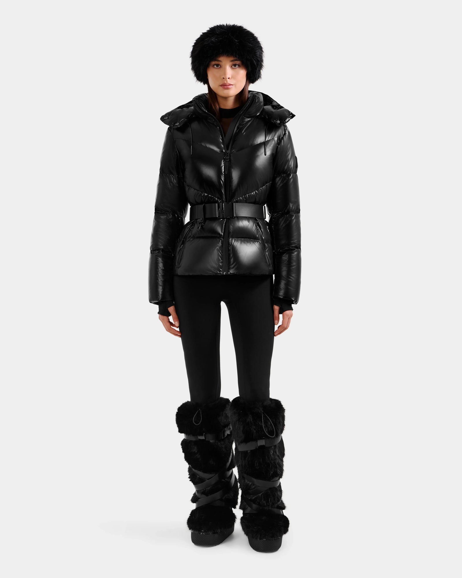 A woman faces forward in a shiny black Rudsak CHRISTELLE S - L125544 down puffer jacket with a belt and faux fur trim, paired with black leggings, furry boots, and a matching furry hat against a plain light gray background.