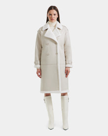 A woman faces forward in the Rudsak DARCY - L125539 Vanilla long, light beige winter coat with wide lapels and double-breasted buttons, paired with a white turtleneck and knee-high white boots against a plain white background.