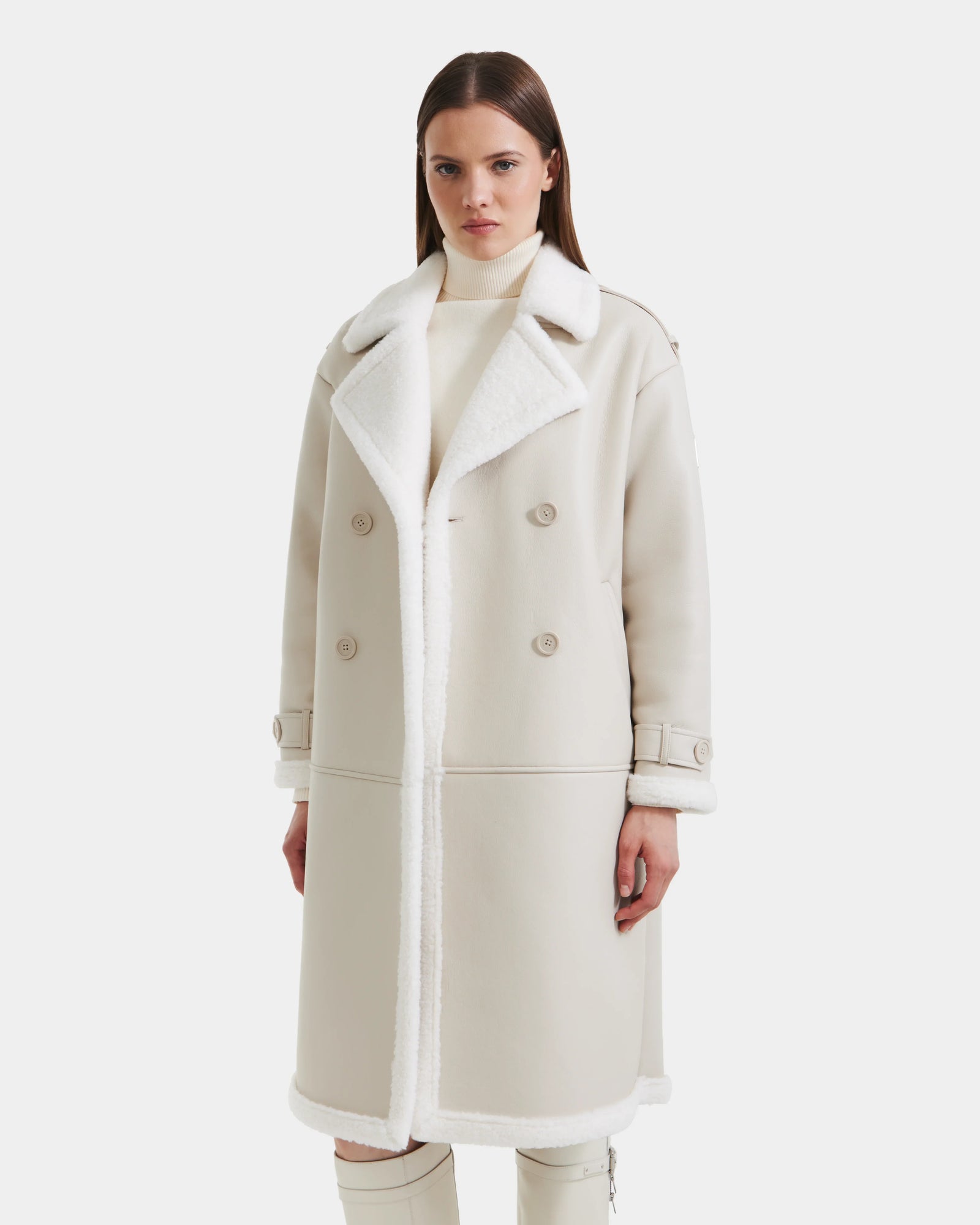 A woman stands facing forward, wearing the Rudsak DARCY - L125539 Vanilla long faux sherpa coat with wide lapels and double-breasted buttons over a light turtleneck, set against a plain white background.