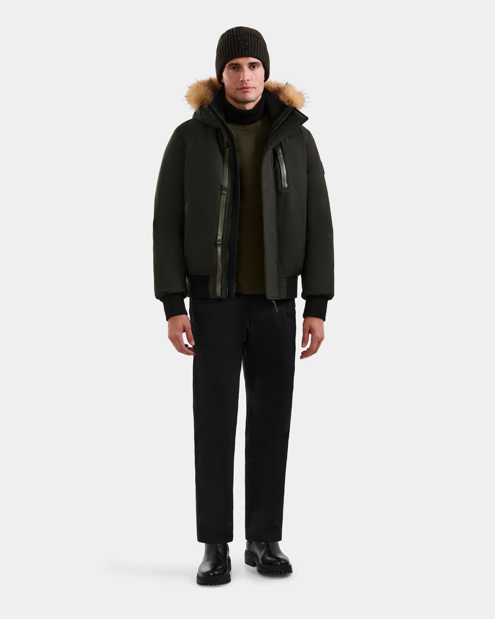 A man faces forward against a plain light background wearing the Rudsak DEACON - M123513 Dark Olive bomber-style down puffer with a fur-lined hood, styled with black pants, black boots, a dark green sweater, and a dark knit beanie.