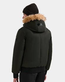 A person wearing the Rudsak DEACON - M123513 men's down puffer coat in dark olive with light brown fur trim, a black knit beanie, and black pants, shown from the back against a plain light background.