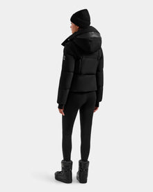 A person wearing a Rudsak DELILAH M - L125538 Black women’s down puffer with removable sleeves, black leggings, winter boots, and a black beanie stands facing away from the camera against a plain light gray background.