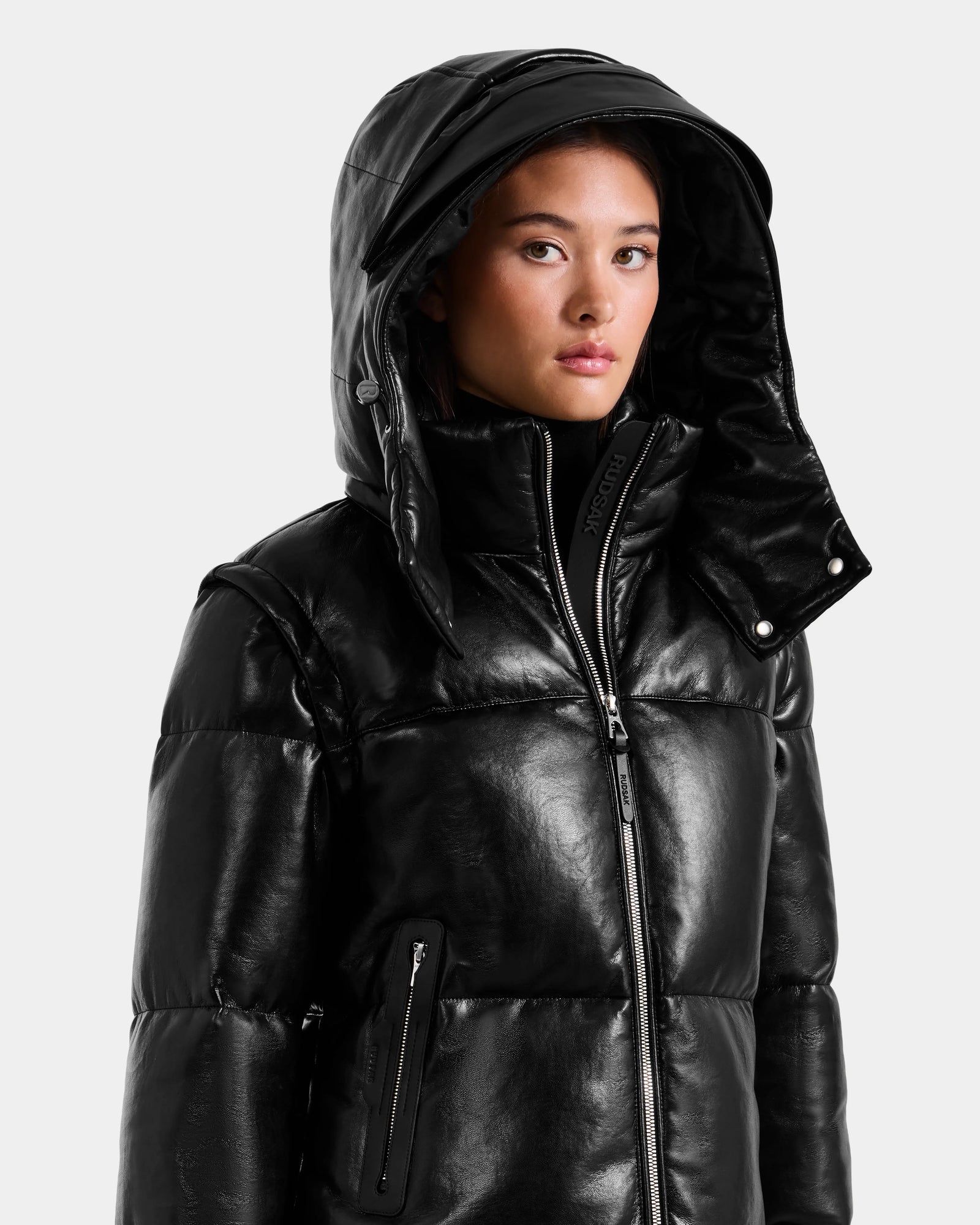 A person wears the Rudsak DELILAH PX - L125537 Black women’s vegan-leather puffer jacket, featuring a large hood, high collar, and visible zippers. The figure faces slightly sideways against a plain light backdrop.