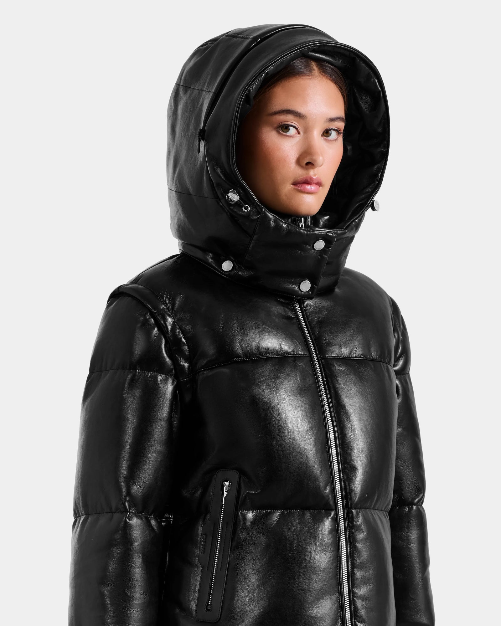 A young woman wears the Rudsak DELILAH PX - L125537 black vegan-leather puffer jacket with a high zip-up collar and hood, looking slightly to the side against a plain, light background.