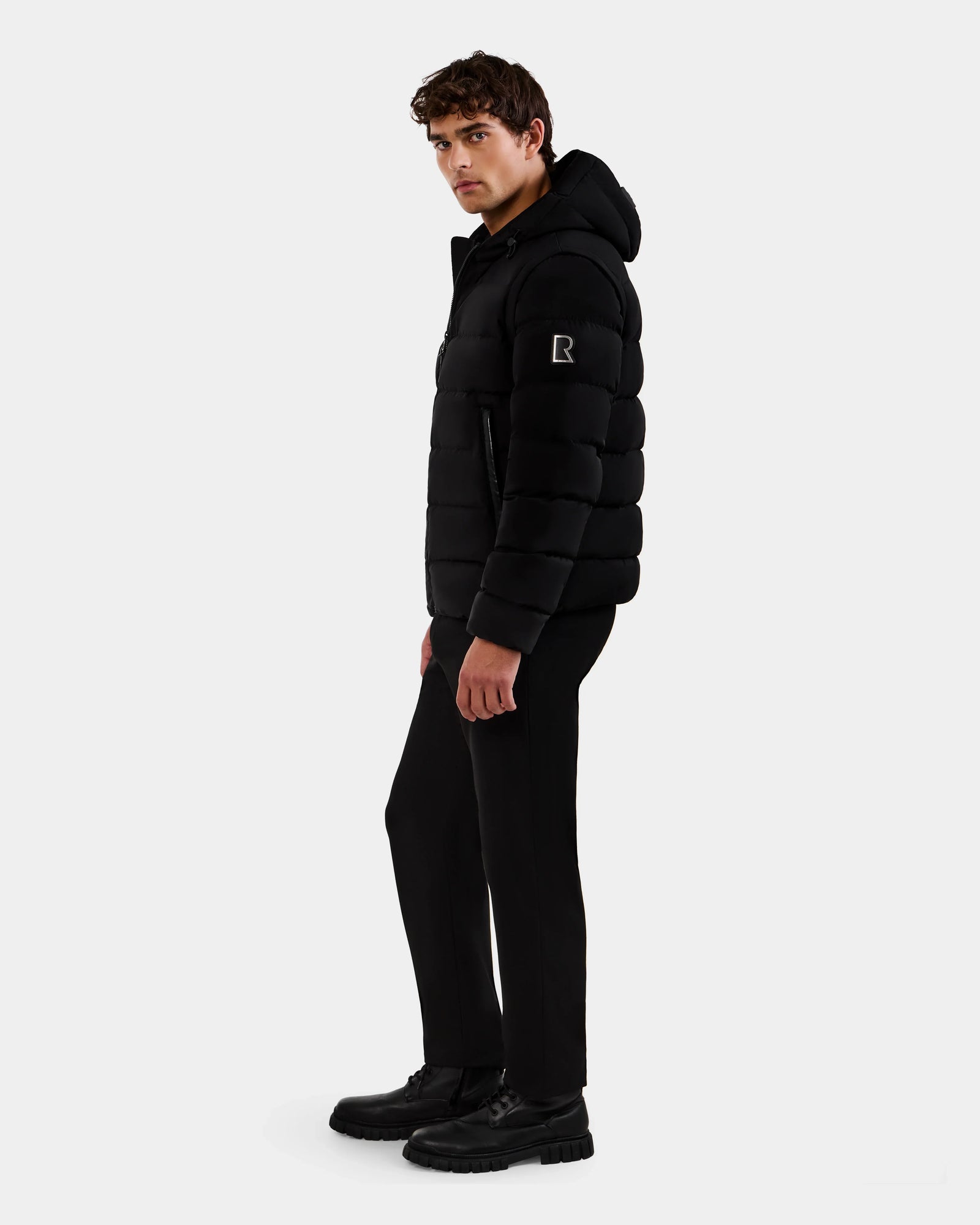 A man stands sideways against a plain light background, wearing the Rudsak DEMIAN M-R - M123504 Black men's down puffer jacket with black pants and black shoes.