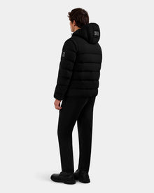 A person wearing a Rudsak DEMIAN M-R - M123504 Black men's down puffer jacket, black pants, and black shoes stands facing away from the camera against a plain light gray background.
