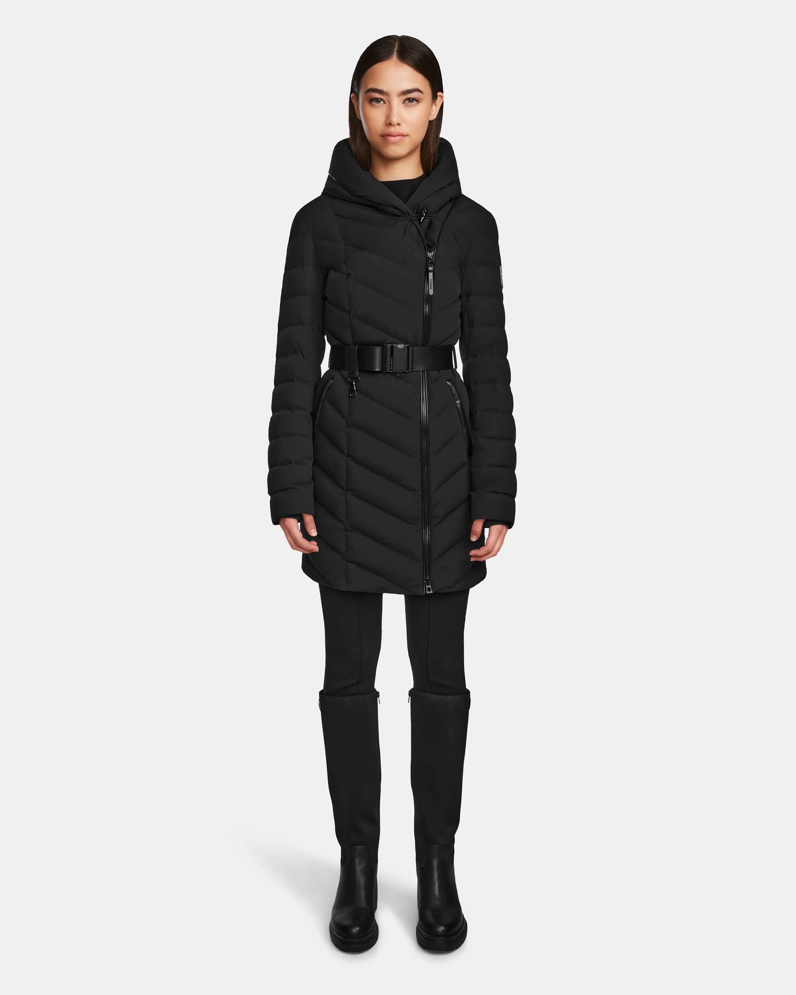 A woman faces forward wearing the Rudsak DITA - L124556 Black knee-length puffer jacket with a high collar and zippered front, paired with black pants and knee-high boots against a plain white background.