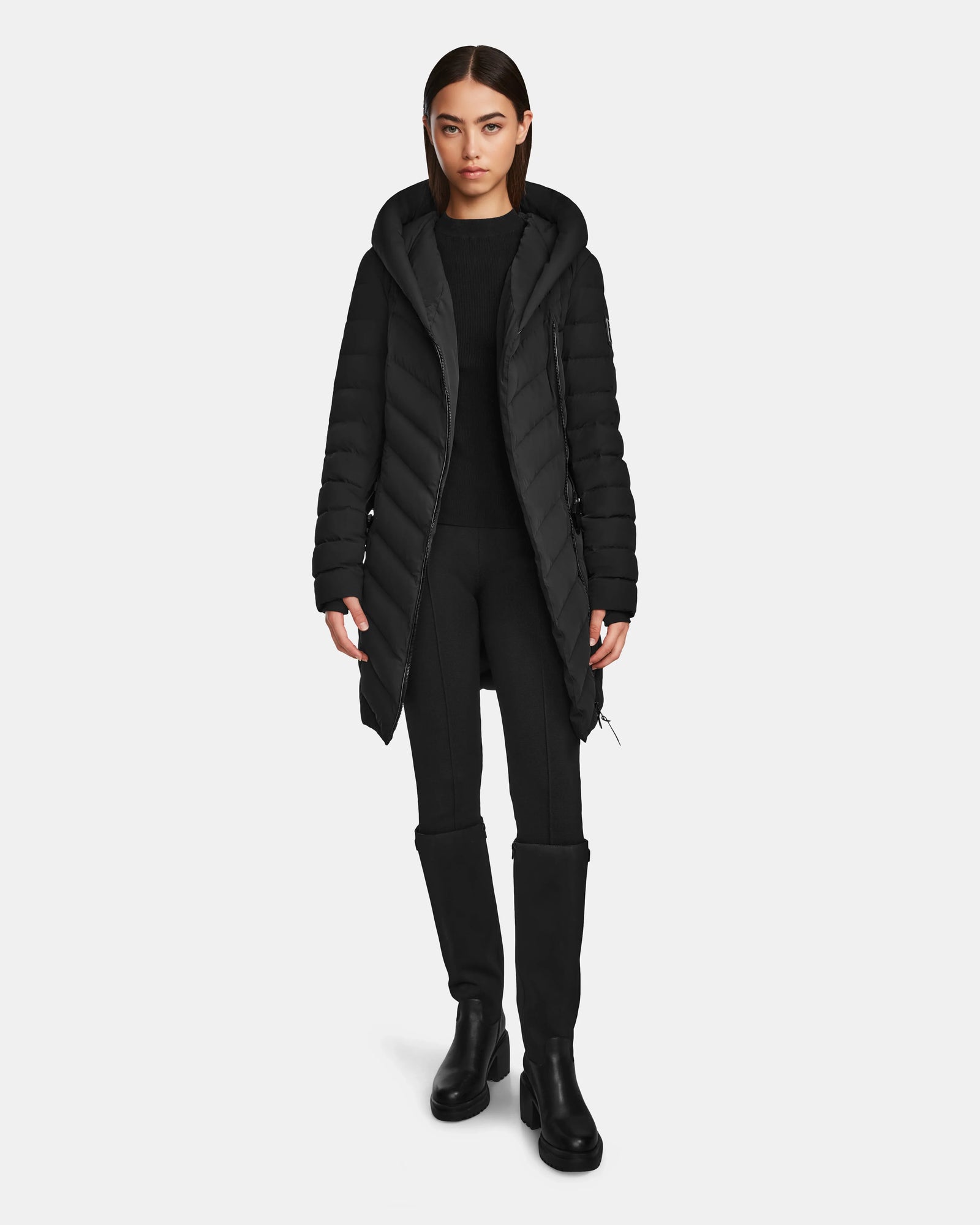 A woman stands facing forward in the Rudsak DITA - L124556 Black knee-length puffer jacket with a hood, black pants, and boots. Her straight brown hair frames her neutral expression against a plain white background.