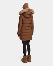 A person wearing the Rudsak ELSA - L123535 Taupe womens quilted down puffer coat with a fur-lined hood faces away from the camera, paired with black leggings, brown boots, and a brown knit beanie against a plain background.