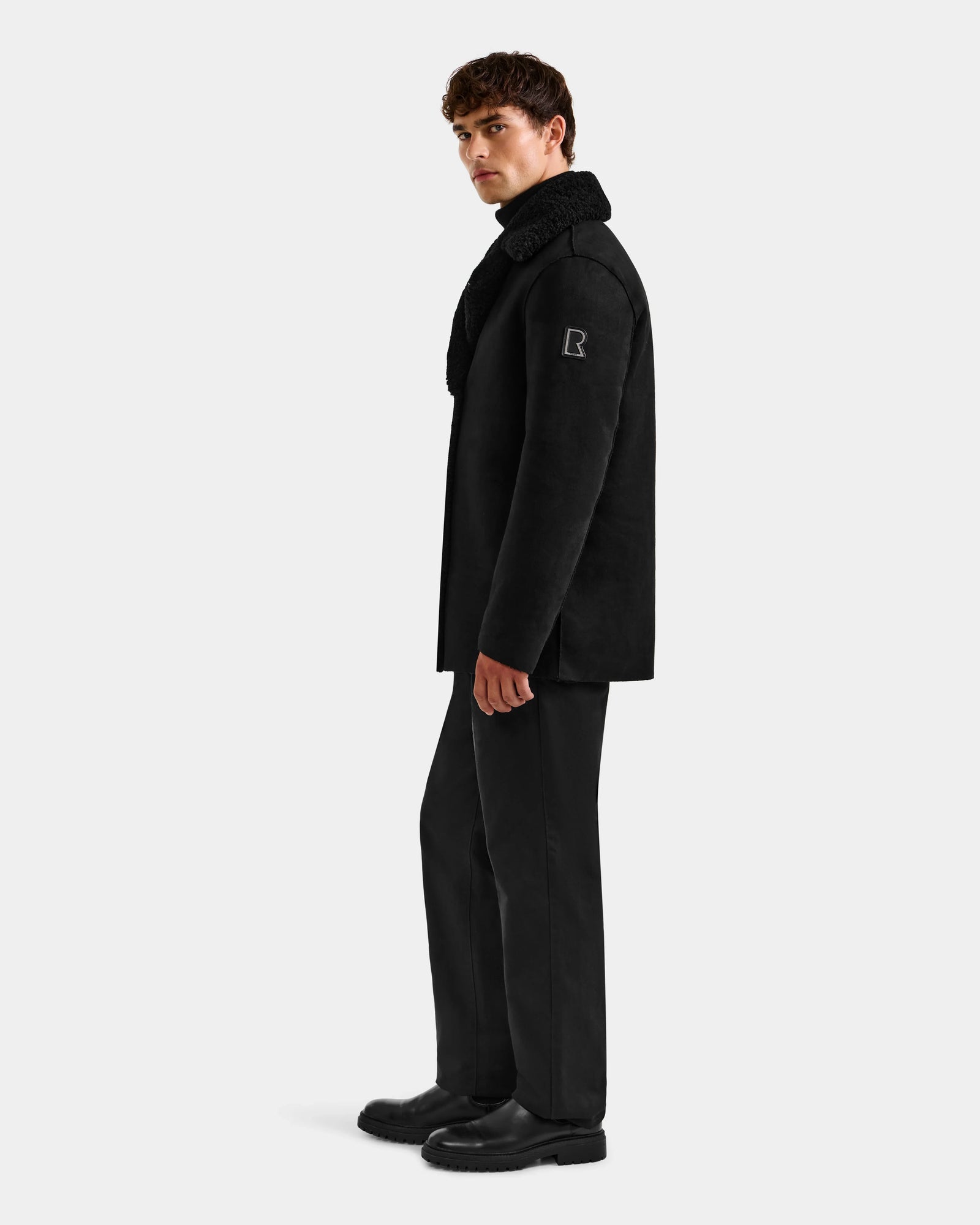 A man stands sideways against a plain light background, wearing the Rudsak ELTON - M125534 Black faux shearling coat, paired with black pants and black shoes.