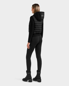 A woman faces away, wearing the Rudsak EMILY - L125515 Black women’s hooded down bomber vest with RDS-certified down, black long-sleeve top, black leggings, and lace-up boots, against a plain light gray background.