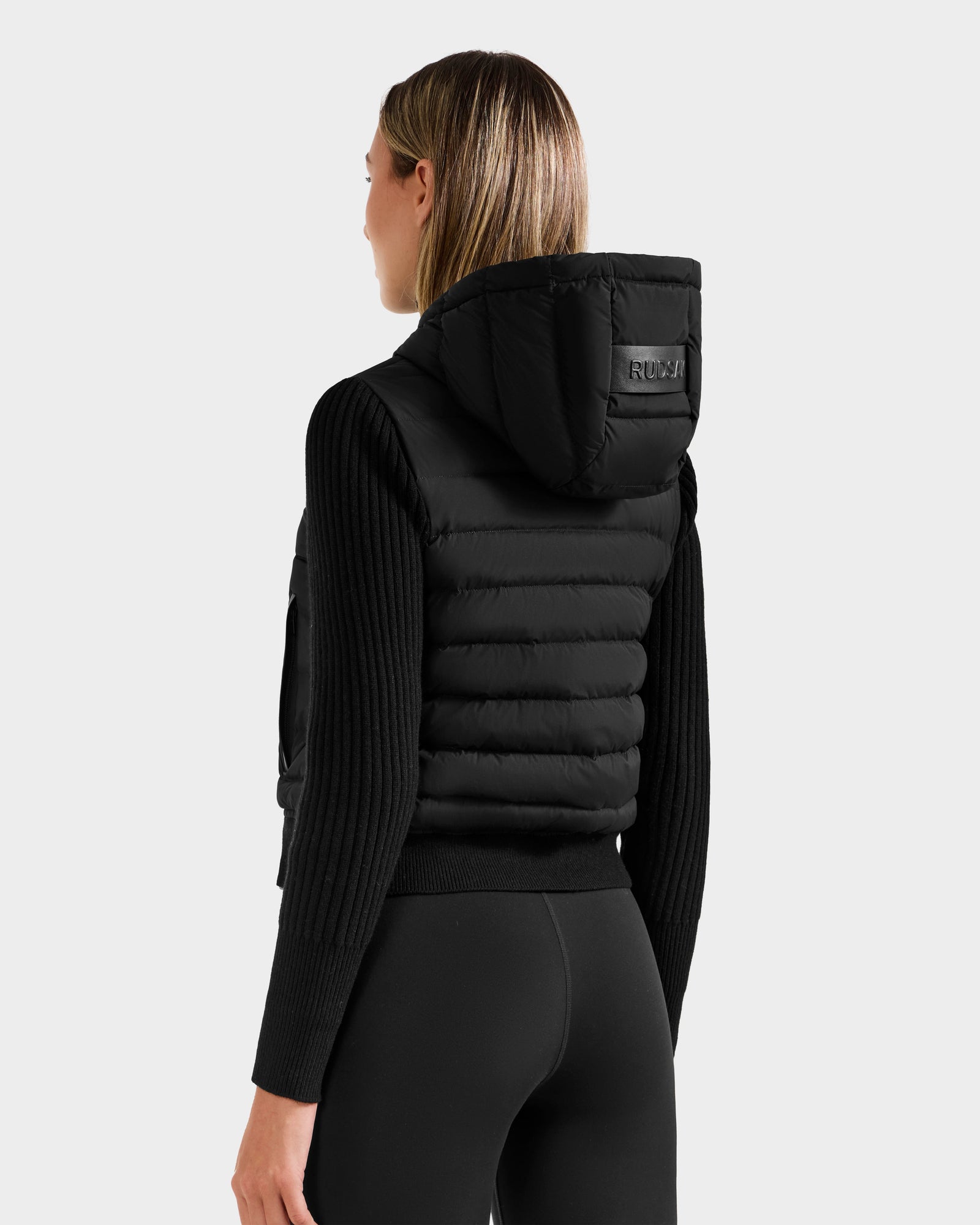A woman with straight, shoulder-length hair is seen from behind wearing the Rudsak EMILY - L125515 Black women’s hooded down bomber, featuring a logo patch on the hood and filled with RDS-certified down for warmth.