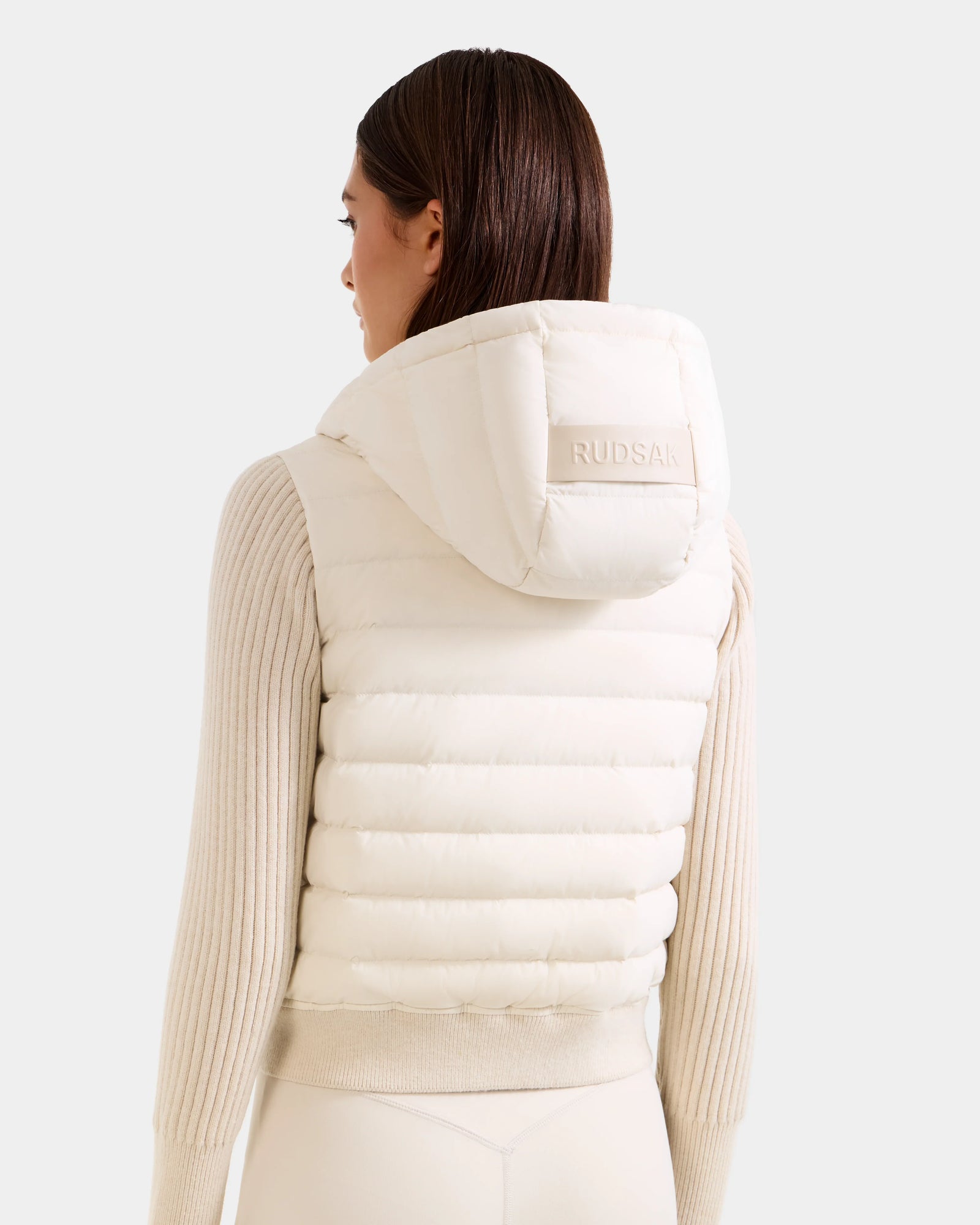 A woman with straight brown hair wears the Rudsak EMILY - L125515 Vanilla women’s hooded down bomber vest, embroidered with RUDSAK on the hood, over a ribbed long-sleeve sweater, standing against a light plain background.