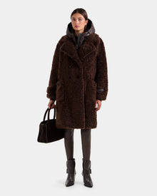 A woman with straight brown hair and a neutral expression wears the Rudsak EMRYN - L125588 Espresso long brown faux-fur winter coat over a black puffer jacket, brown leggings, black ankle boots, and holds a black handbag.