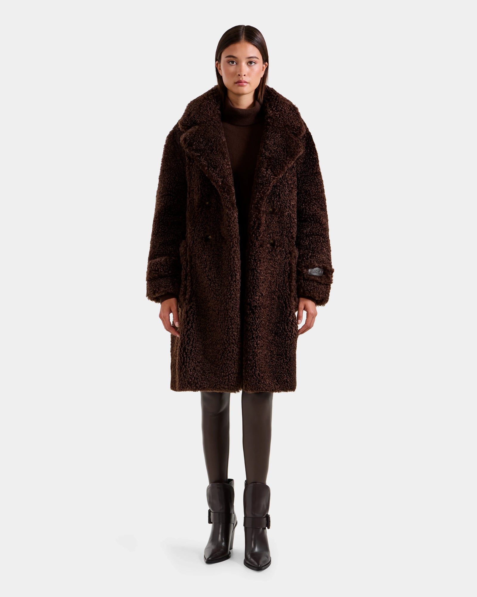 A woman faces forward against a plain light background, wearing the Rudsak EMRYN - L125588 Espresso double-breasted teddy jacket over a brown top, black leggings, and black buckled ankle boots.