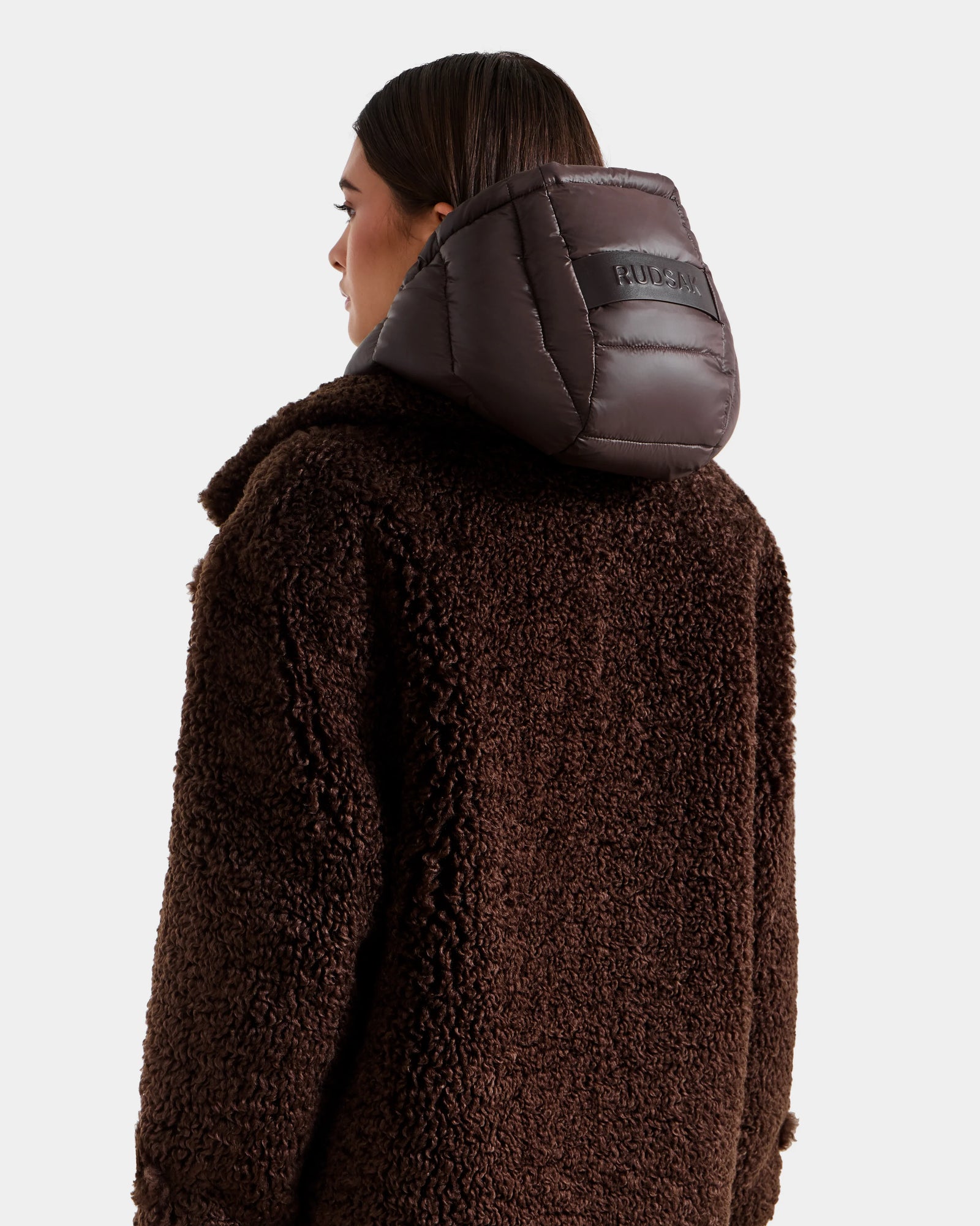 A woman wears the Rudsak EMRYN - L125588 Espresso women’s teddy jacket with a textured finish and shiny padded hood, shown from the back against a plain light background.