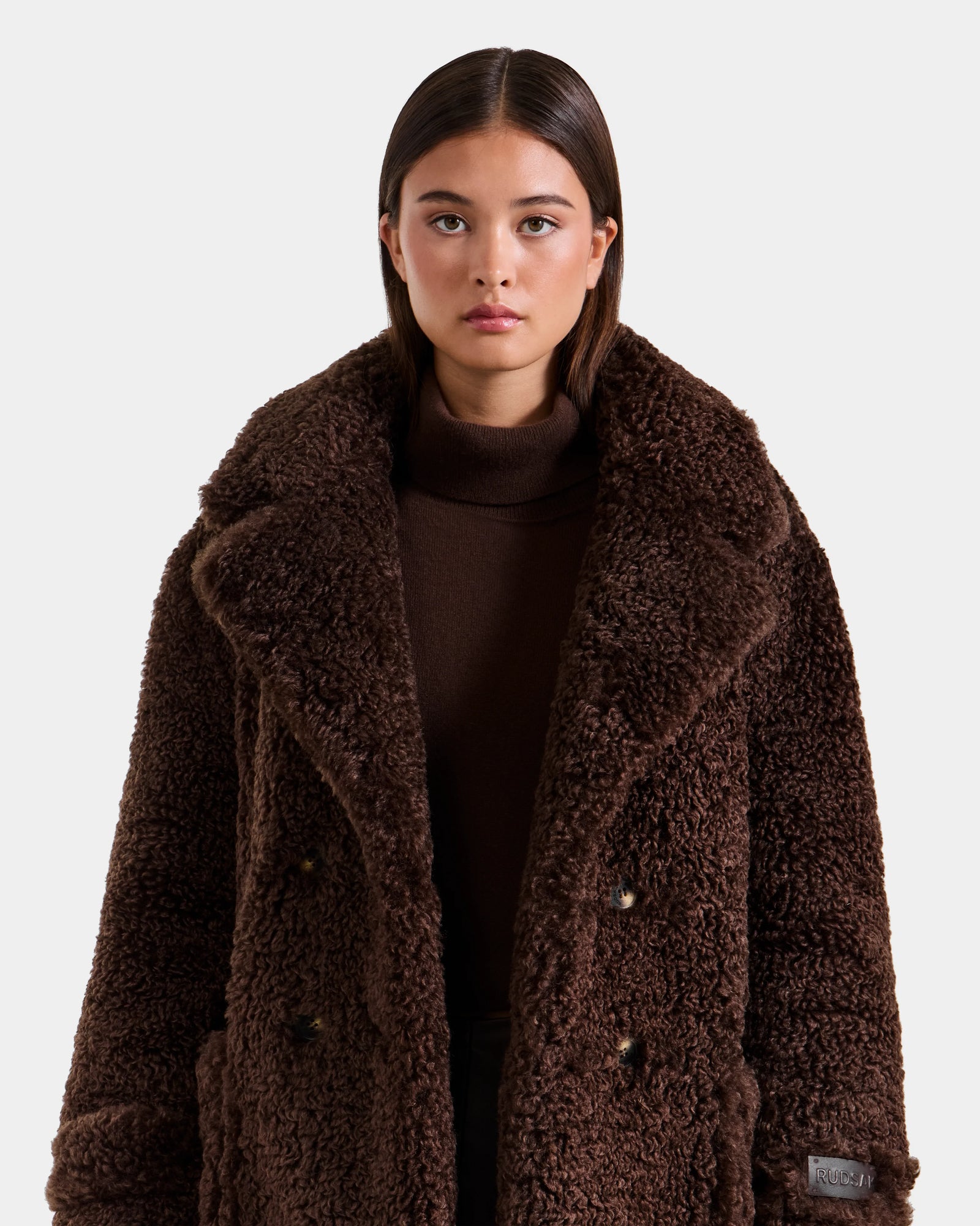 A woman with straight brown hair wears Rudsak’s EMRYN - L125588 Espresso teddy jacket over a brown turtleneck, standing against a plain, light background.