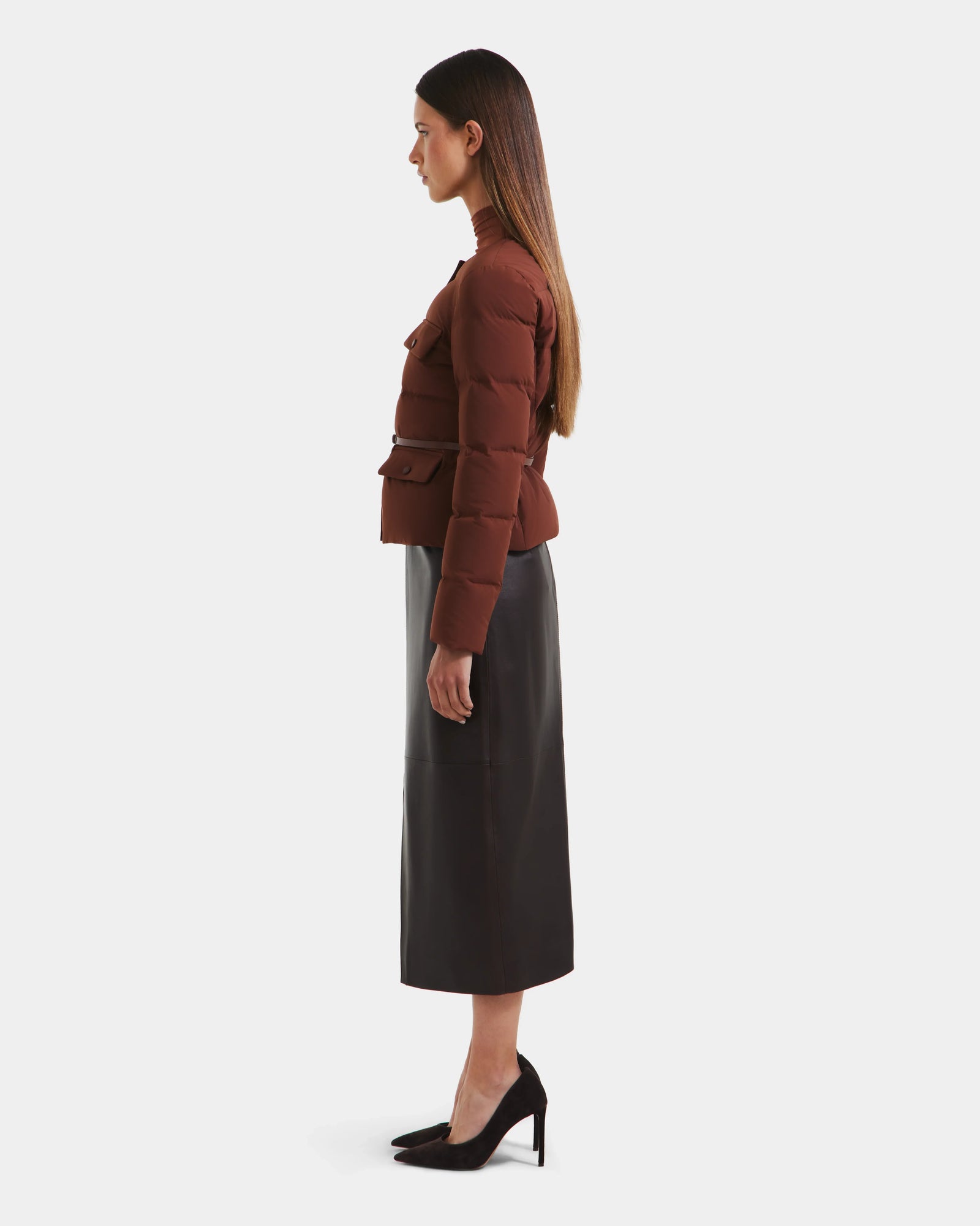 A woman with straight brown hair stands in profile, wearing the Rudsak FRANCESCA - 8125021 Rust quilted puffer jacket, a black midi skirt, and black high heels against a plain white background.