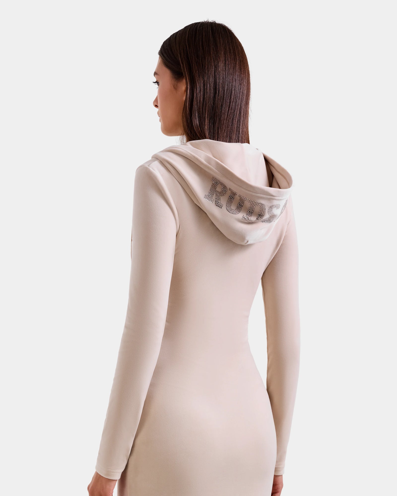 A woman with straight brown hair models the Rudsak ISABELLA - L525502 Cream, a fitted, long-sleeve beige velour hooded midi dress. "Rude" in silver sequins accents the back as she looks away against a plain background.