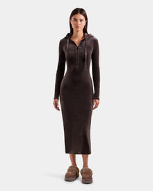 A woman stands facing forward, wearing the Rudsak ISABELLA - L525502 Espresso women’s velour hooded dress with a front zipper and fitted long sleeves, paired with tan fuzzy slippers, on a plain light background.