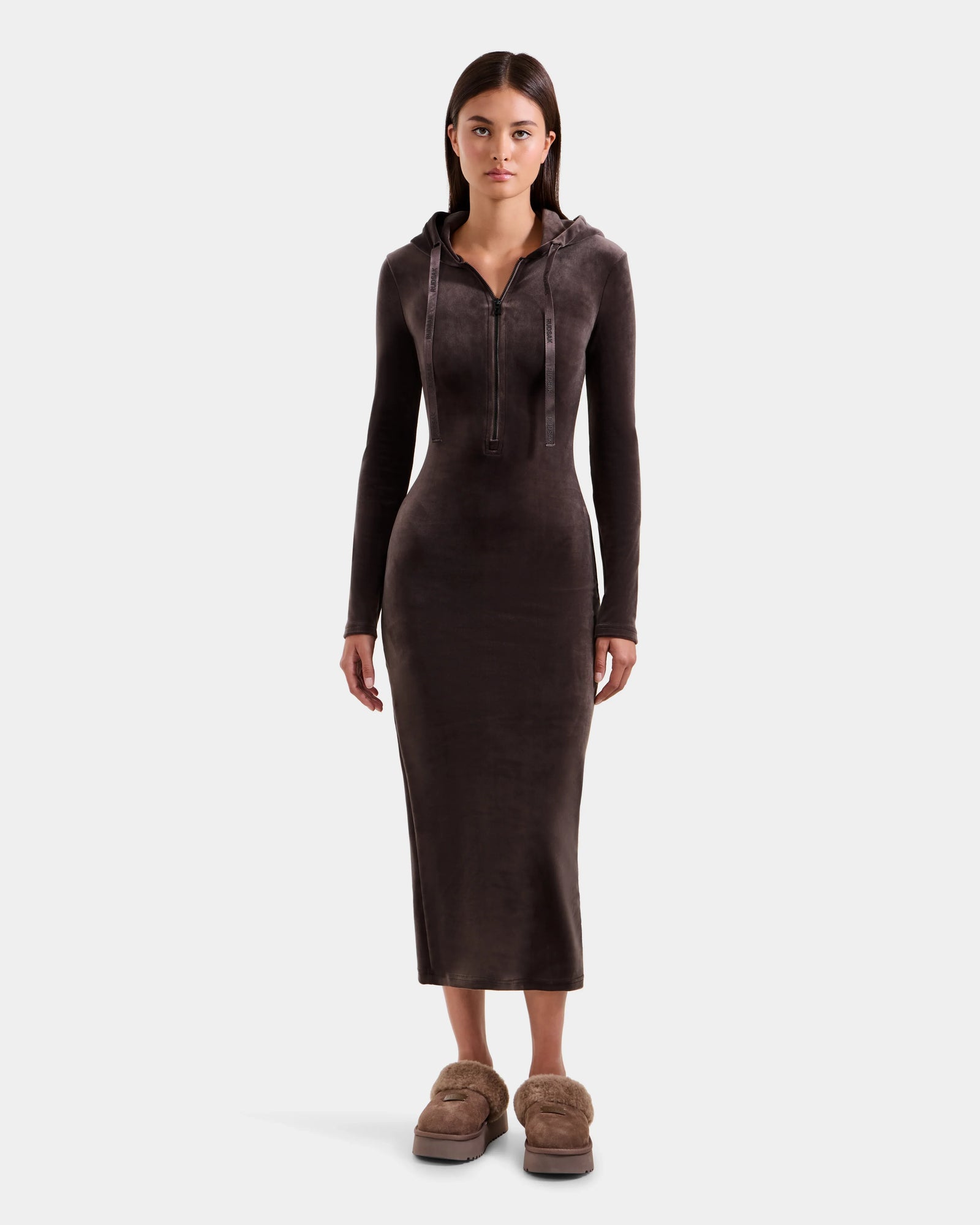 A woman stands facing forward, wearing the Rudsak ISABELLA - L525502 Espresso women’s velour hooded dress with a front zipper and fitted long sleeves, paired with tan fuzzy slippers, on a plain light background.