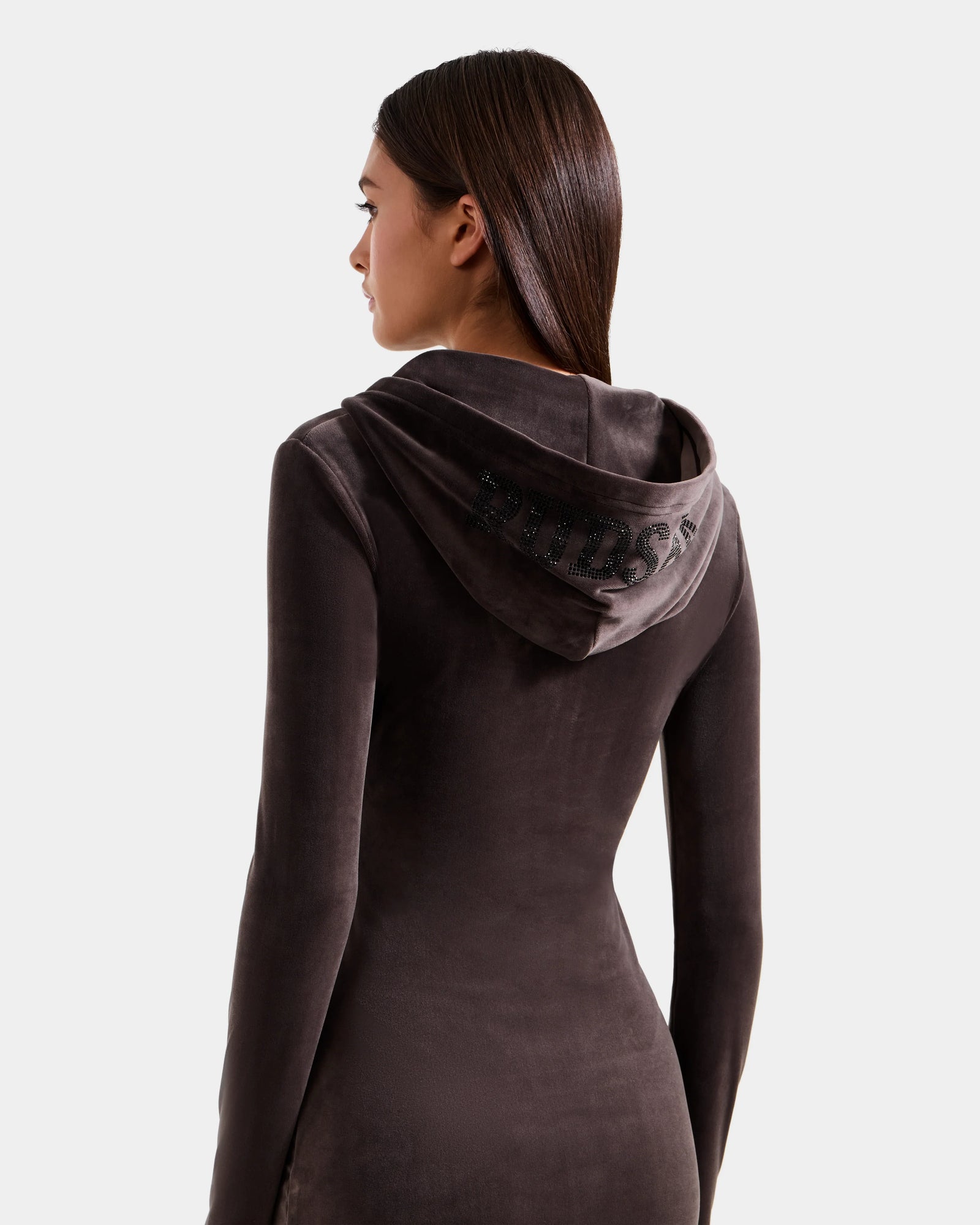 A woman with straight brown hair, seen from the back, wears the Rudsak ISABELLA - L525502 Espresso women’s velour hooded dress featuring rhinestone "PRINCESS" on the hood. The background is plain light gray.