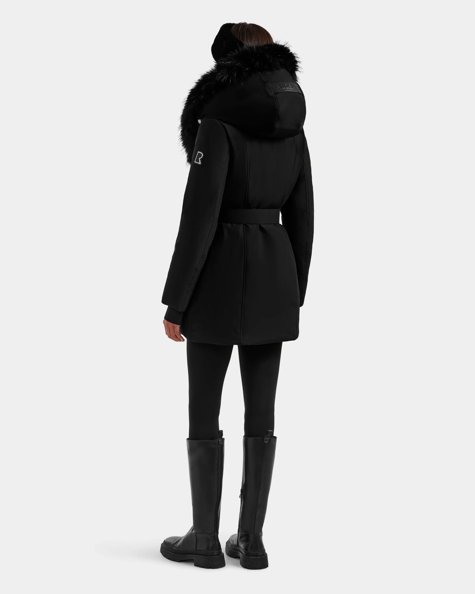 A person stands with their back to the camera against a plain light gray background, wearing the Rudsak ISELLE - L125547 Black women's down parka with fur trim, paired with black leggings and knee-high boots.
