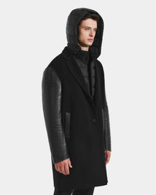 A man wears the Rudsak JESSE - M125504 Black men’s wool coat with leather sleeves and a shiny, down-insulated hood, standing against a plain white background.
