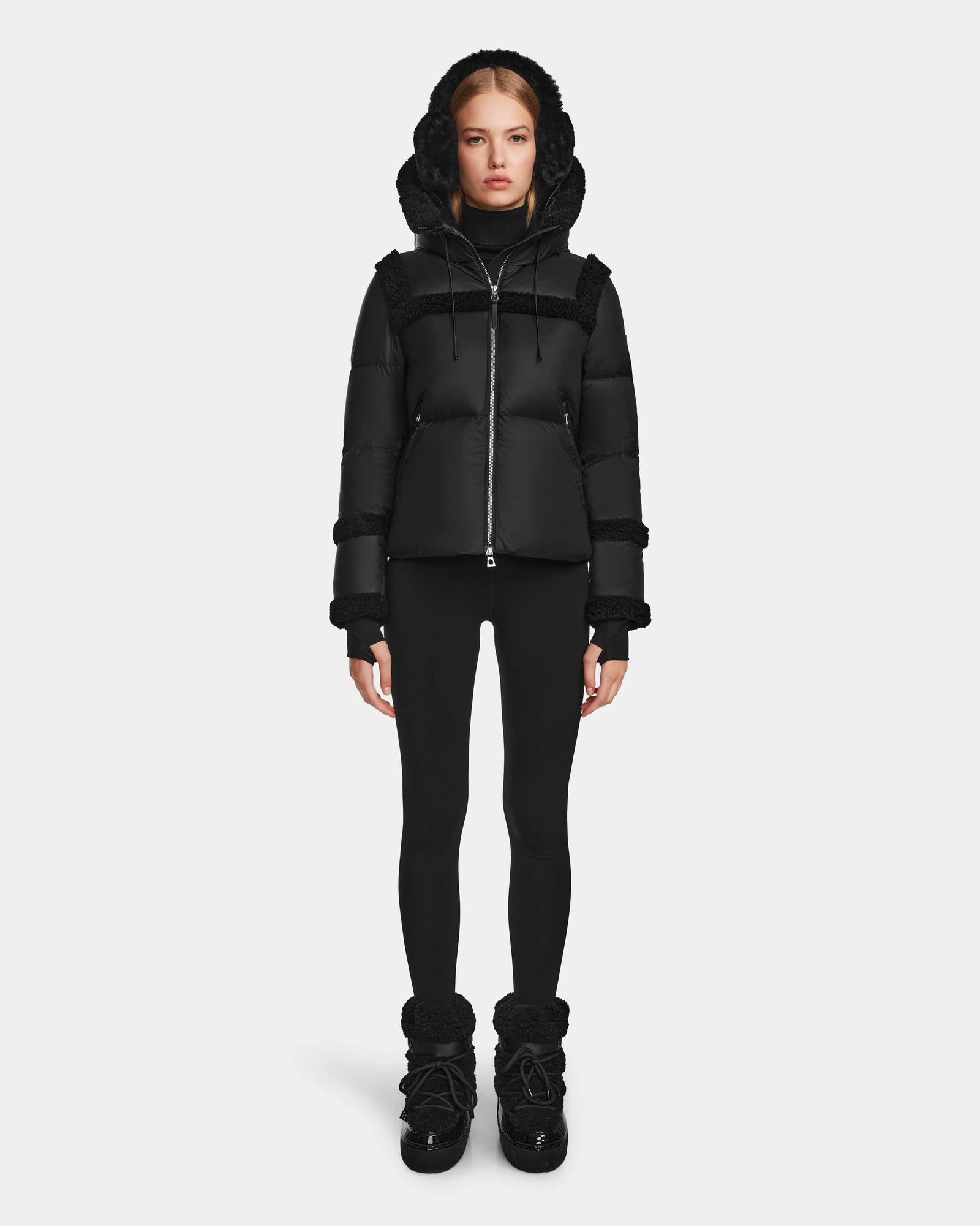 A woman stands facing forward against a plain white background wearing the Rudsak JULIANA - L124509 Black/Black women's short down puffer with a hood, black leggings, and black winter boots.