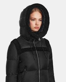 A woman wears the Rudsak JULIANA - L124509 Black/Black short down puffer with a black fleece-lined hood, looking straight ahead against a plain light background.