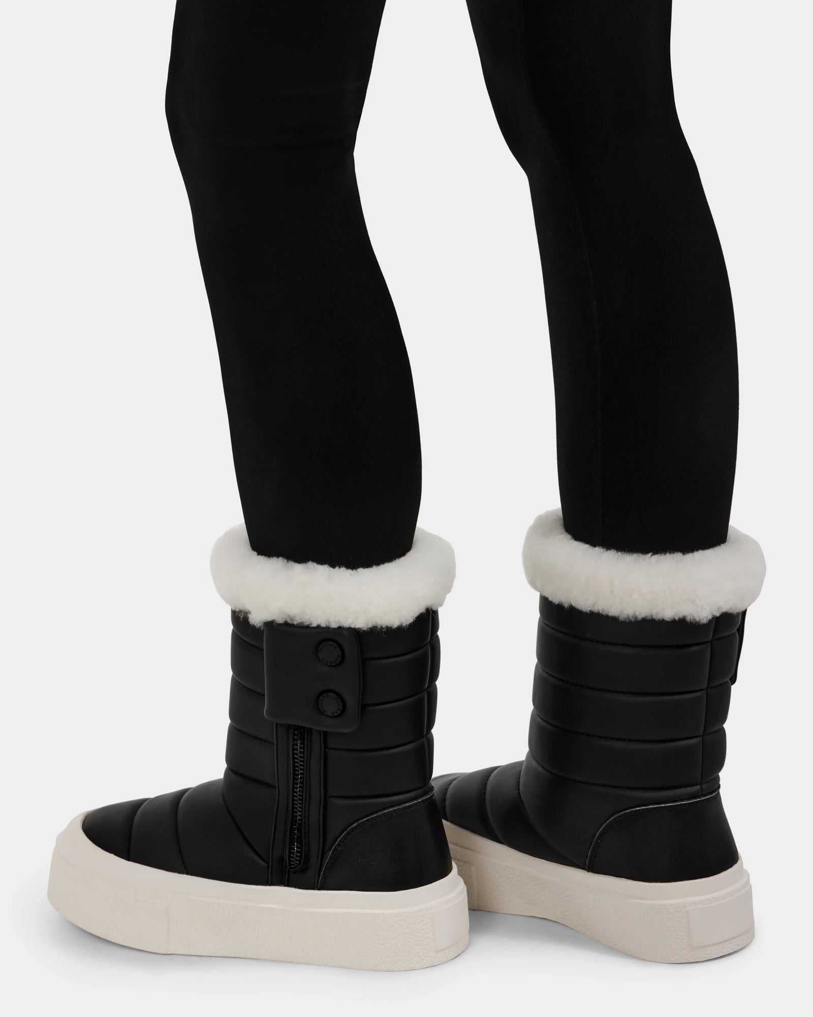 A person wears Rudsak KATHIE - L225516 Black women's quilted insulated mid boots with white faux fur trim, thick white soles, side zippers, and buttons, styled with black leggings for a cozy winter look.