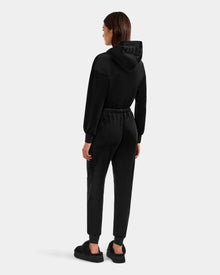 A woman faces away, wearing Rudsak’s KETZY - L525503 Black velour hooded jumper with an elastic waistband and cuffed pants, paired with black platform shoes, set against a plain light gray background.