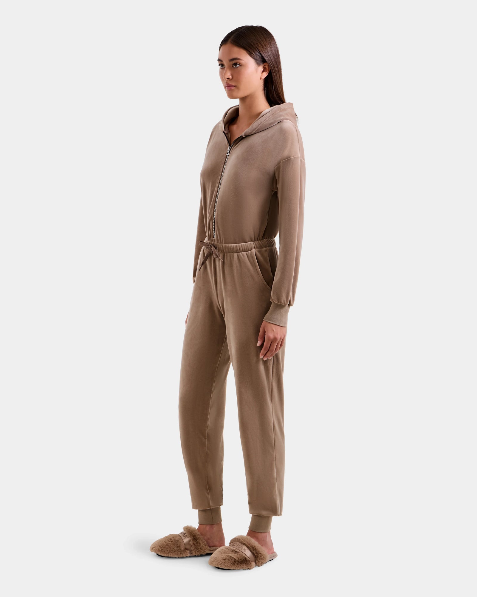 A woman models the Rudsak KETZY - L525503 Sand plush stretch velour zip-up hooded jumpsuit with a drawstring waist and matching fuzzy slippers, facing slightly left against a plain light background.