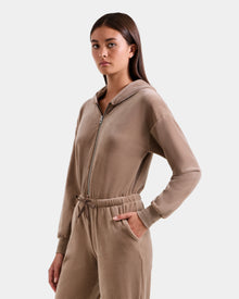 A young woman stands with one hand in her pocket, wearing the Rudsak KETZY - L525503 Sand plush stretch velour zip-up hooded jumpsuit with long sleeves and a drawstring waist, set against a plain light gray background.