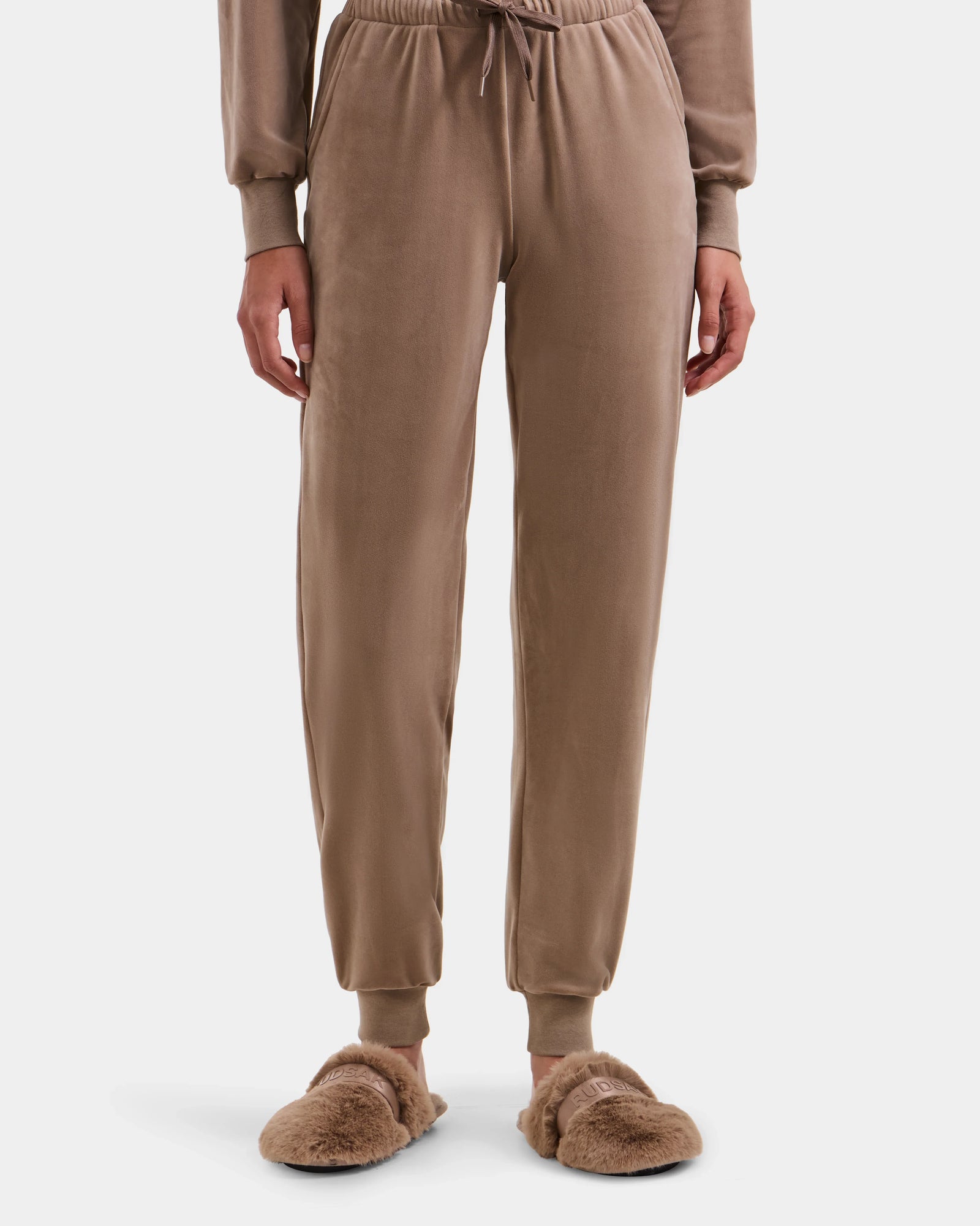 Wearing the Rudsak KETZY - L525503 Sand velvet jogger pants with cuffed ankles and brown furry slippers, paired with a women’s plush stretch velour hooded jumper, a person stands against a plain white background.