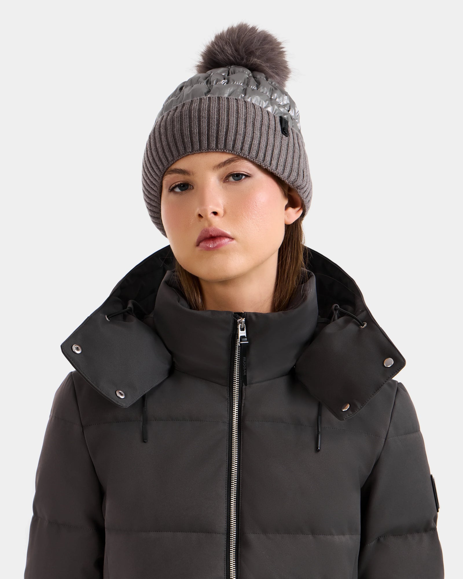 Wearing the Rudsak LANYA - L423514 Pewter puffer jacket, a woman with a high collar and a women’s pom pom hat gazes ahead with a neutral expression against a simple light background.