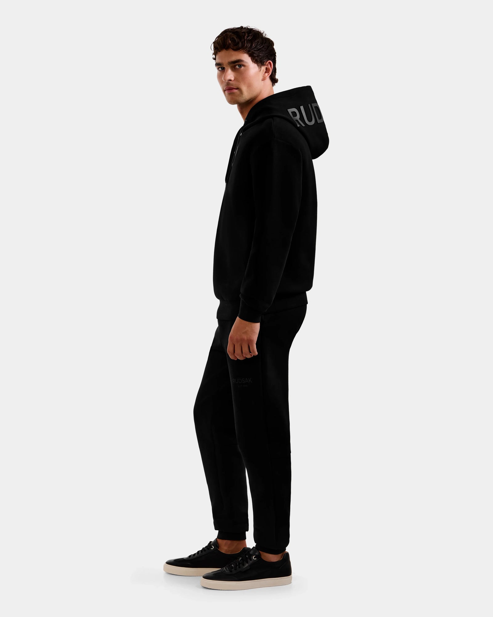 A young man models the Rudsak LEROY - U525509 Black M unisex hoodie with joggers, both in black luxe cotton-blend, paired with black sneakers featuring white soles, set against a plain light background.